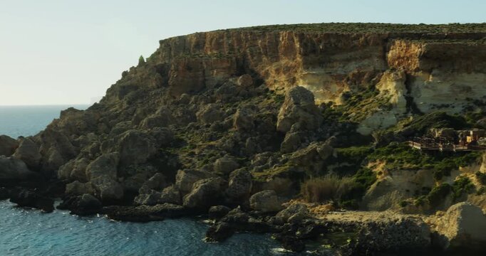 Steep grassy and rocky seashore with famous small village on island of Malta at sunset. Buildings of Popeye settlement scattered on slope opposite bay