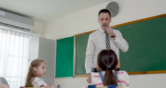 Young blond boy raises hand from wooden desk to answer teacher question while multicultural classmates listen, creating interactive elementary classroom discussion during first day school routine.