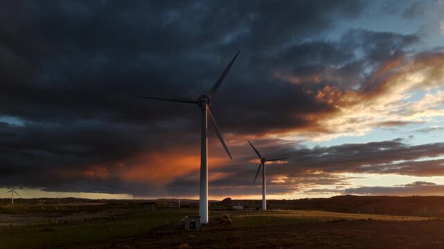 Drone footage of wind turbines on rural moorland with dark storm clouds and orange sunset light in Scotland.