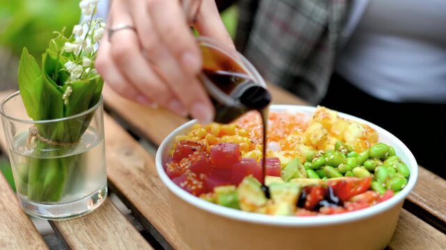 Woman hands pouring soy sauce on healthy tuna poke bowl at wooden table with lily of valley flowers.