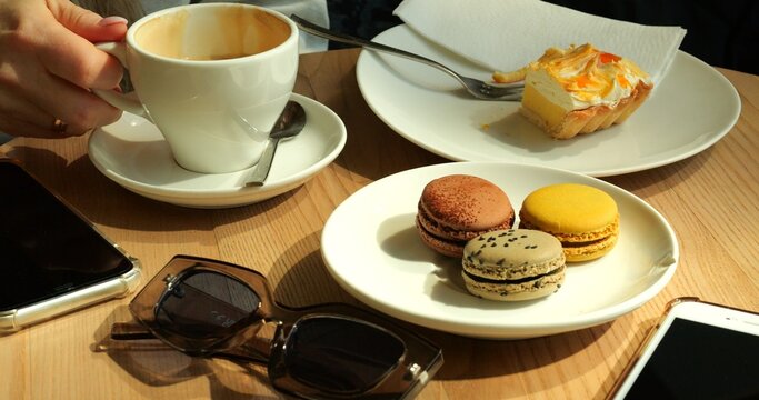 Woman sips coffee savoring assortment of macarons and luscious slice of pastry. Smartphones and stylish sunglasses casually put on table near plate