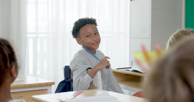 Young boy student turns from wooden desk toward male teacher during primary classroom discussion, joining diverse children learning together through first day routine, notebook work and question time