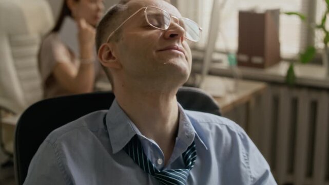 Tilt up shot of Caucasian man in formalwear resting in computer chair, enjoying fresh breeze while using portable mini fan