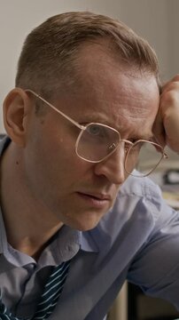 Vertical shot of Caucasian man taking off glasses and sweltering while working on computer in stuffy office