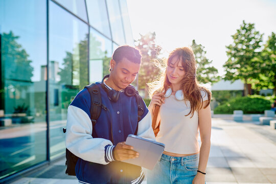 Young diverse students collaborating, sharing ideas on a digital tablet, walking outdoors at university campus on a sunny day