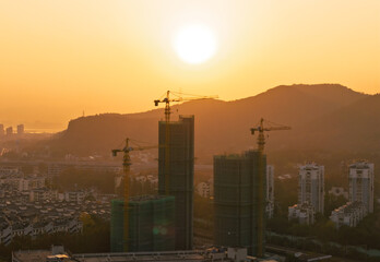 Fototapeta premium Building cranes silhouette against a vibrant golden sunset sky over a developing city nestled among distant mountains