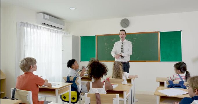 Elementary boy raises arm to answer teacher question while male educator stands near chalkboard, discussion builds confidence, participation, discipline, active learning during school opening