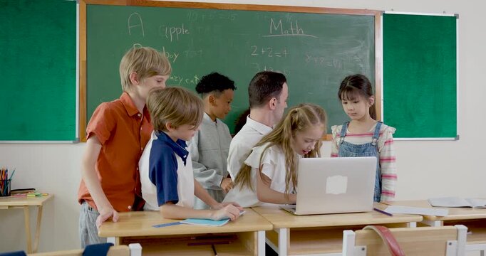 Slow motion group activity with male teacher and multicultural elementary children gathered around laptop, sharing ideas, classwork, teamwork, and first day school excitement near chalkboard.