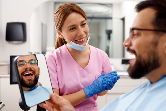 Smiling male patient looking at his reflection in mirror while female dentist explains treatment results in modern dental clinic