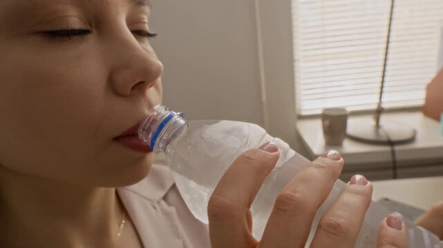 Tracking shot of Caucasian woman sitting at work desk, opening bottle of cold water and drinking slowly