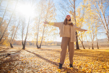 Woman standing in autumn park with arms outstretched enjoying sunny day. Happy girl in warm sweaters walking with dog in forest. Nature landscape for seasonal lifestyle concept. © Tatyana Gladskih