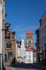 Obraz premium A narrow old town street leads toward a brick church tower and red roofs in Rostock, Mecklenburg-Western Pomerania, Germany.
