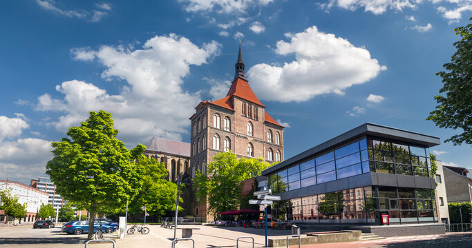 A historic brick building and modern glass facade stand together on a sunny street in Rostock, Mecklenburg-Western Pomerania, Germany.