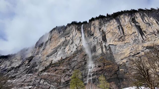 Slow tilt down of Staubbach Falls in Lauterbrunnen, Switzerland, as snow falls across towering cliffs and the famous waterfall descends into the winter valley.