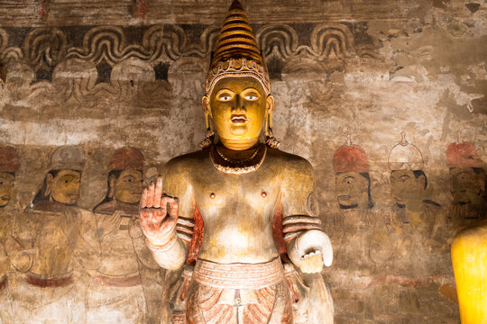 View of a golden Buddha statue with ancient mural paintings on the stone walls inside the historic cave temple in Dambulla, Central Province, Sri Lanka.
