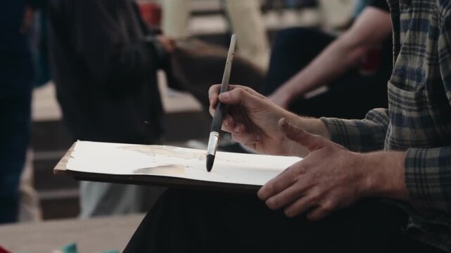 Creative painter's hands holding a brush and working on a watercolor painting outdoors, capturing a moment of artistic expression and skill in a public space with a shallow depth of field