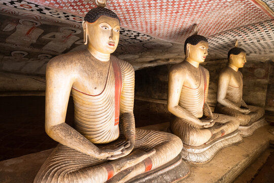 View of ancient seated Buddha statues with saffron robes and intricate ceiling murals inside the Dambulla Cave Temple Dambulla, Central Province, Sri Lanka.