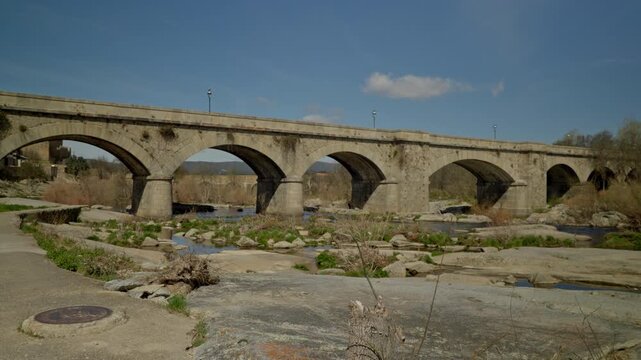 Ancient stone arch bridge crossing a low water river with rocks and dry veget...