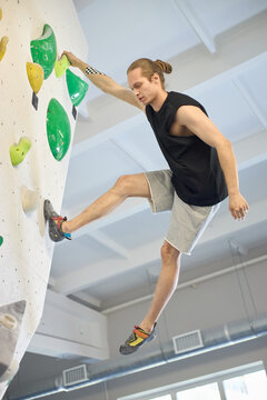 Vertical shot of young adult Caucasian man climbing indoor bouldering wall, training strength and focus for fitness challenge