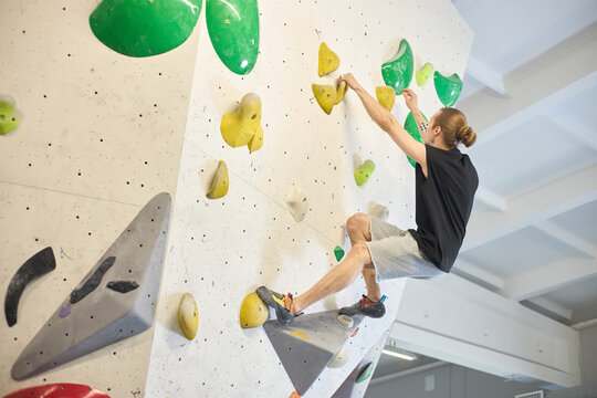 Young adult Caucasian man climbing indoor bouldering wall, reaching holds and balancing on footholds, showing fitness challenge and active lifestyle
