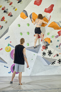 Vertical shot of young adult woman climbing indoor bouldering wall while young adult man spotting below, showing fitness training and teamwork