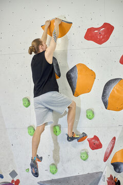 Caucasian young adult man climbing indoor bouldering wall, gripping holds and stepping upward during fitness training