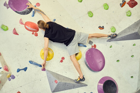 Overhead shot of young adult Caucasian man climbing indoor bouldering wall, stretching across holds and volumes, showing fitness challenge