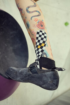 Close up of young adult climber resting tattooed leg in climbing shoe on gym hold, showing kinesiology tape for ankle support