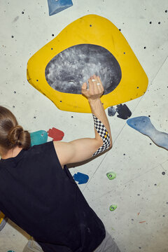 Vertical shot of young adult man climbing indoor bouldering wall, gripping hold and training upper body strength