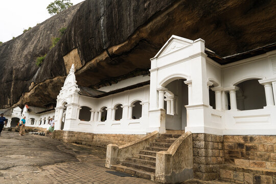 View of the Dambulla Cave Temple featuring white arched entrances built into a massive rock overhang with tourists walking on the stone path Dambulla, Central Province, Sri Lanka.