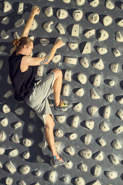 Vertical shot of young adult Caucasian man climbing indoor bouldering wall, gripping holds and training strength, focus, endurance