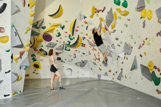 Wide shot of young adult man climbing indoor bouldering wall while young adult woman spotting below, showing fitness training and active lifestyle