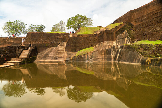 View of the ancient water tank and stone ruins of Sigiriya Rock Fortress with tourists walking on stairs and reflections on the water Sigiriya, Central Province, Sri Lanka.
