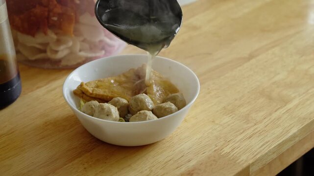 A person pouring hot broth into a bowl containing meatballs and tofu on a dining table setting