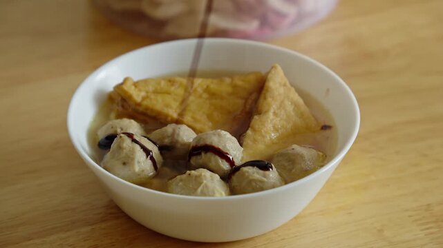 A person pouring sweet soy sauce into a warm bowl of meatball soup on a dining table setting