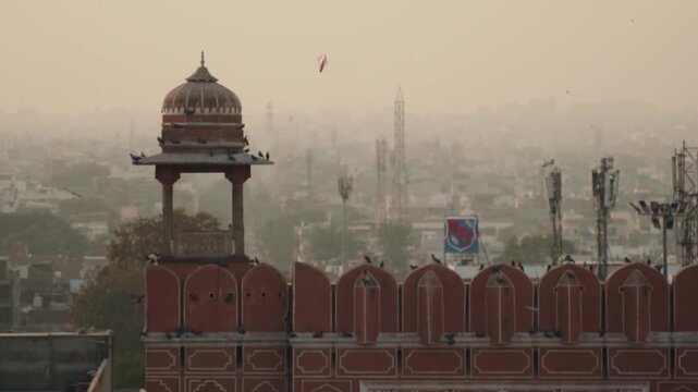 Hazy morning cityscape of Jaipur, India, featuring traditional Rajasthani architecture and a kite flying high in the sky as birds perch on the red building and fly through the air
