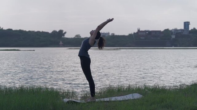 A slow-motion of a South Asian Woman in backbend doing yoga by a calm lake at twilight