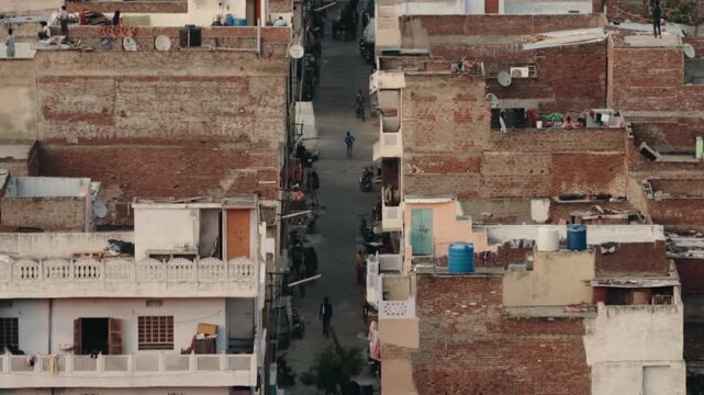 Aerial view of a narrow street in a densely populated Indian city, with people walking and motorcycles parked along the old, weathered brick buildings and rooftops in the warm afternoon light