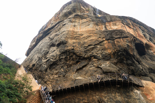 View of the massive Sigiriya Rock Fortress with tourists climbing the metal stairs along the ancient rock face Sigiriya, Central Province, Sri Lanka.