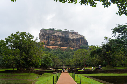 View of the ancient Sigiriya Rock fortress rising above lush gardens and stone ruins with tourists walking along the path in Sigiriya, Central Province, Sri Lanka.