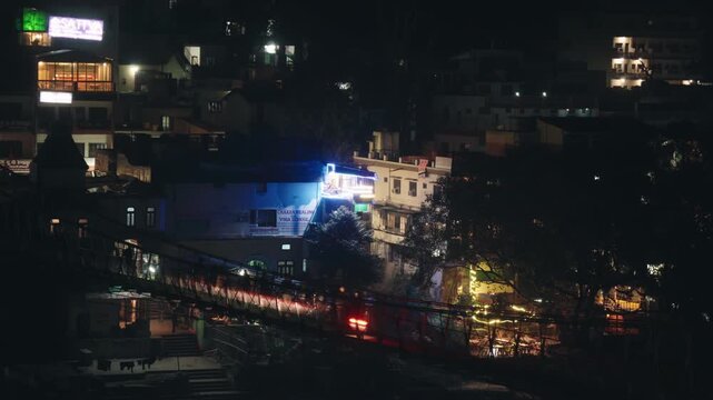 Busy pedestrian traffic crossing the illuminated Laxman Jhula suspension bridge over the Ganges river at night in the holy city of Rishikesh, with city lights and buildings in the background
