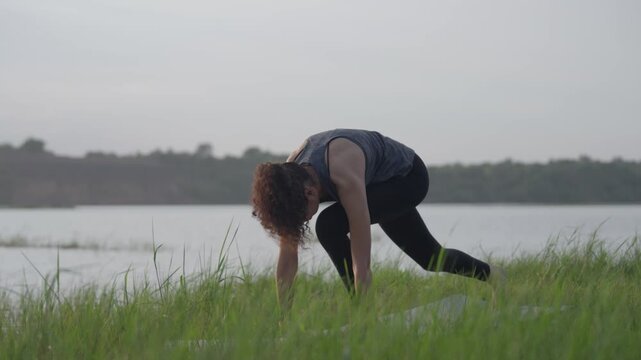 A slow-motion of a South Asian Woman in backbend doing yoga by a calm lake at twilight