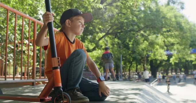 Boy holds scooter upright fixing hat with hand while kneeling in skate park. Child keeps grip on handlebar as fixing blue baseball cap during short riding pause