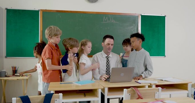 Multicultural elementary student group gathers around male teacher using laptop at desk, discussing class project, sharing notebook work, and building teamwork confidence during school routine.