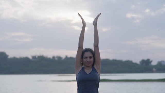 A South Asian Woman in yoga pose with hands clasped overhead, standing by a lake under a pale, cloudy sky