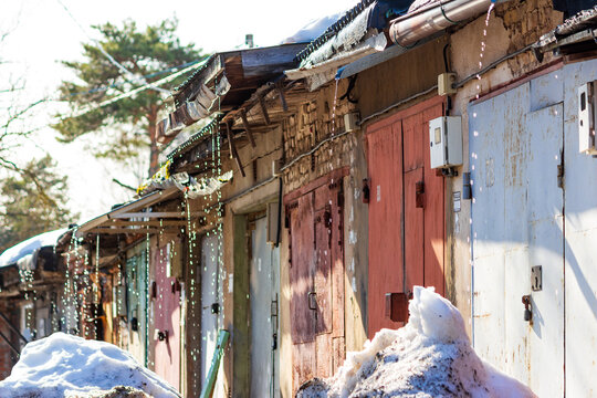 Melting snow dripping from old garage roofs during a bright spring thaw, creating sparkling water streams against the rustic metal doors under the warm seasonal sunlight