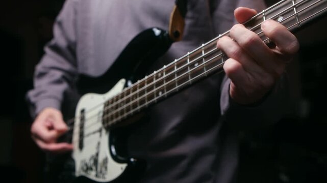 Close up bass player hands plucking and fretting strings black electric bass guitar, rehearsing song in dark studio with focus on musical instrument. Professional musician playing electric bass guitar