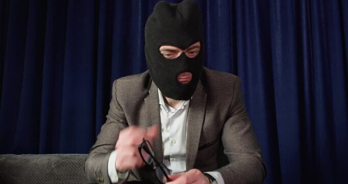 Formerly masked scammer removes balaclava putting on glasses near laptop. Man smiles after disguise change while preparing to continue online fraud work