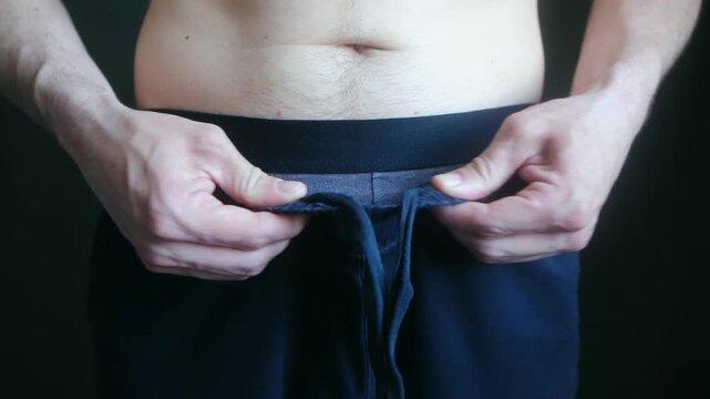 Man struggling to tie his oversized pants after significant weight loss, showcasing the positive results of a diet and healthy lifestyle. His bare abdomen shows the success of his fitness journey