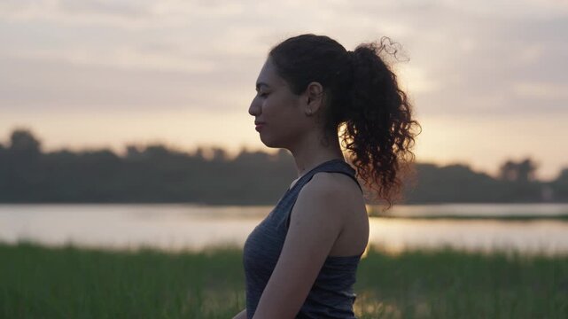 A wide-angle shot of a South Asian woman performing a graceful backbend on her yoga mat beside a quiet lake at sunrise